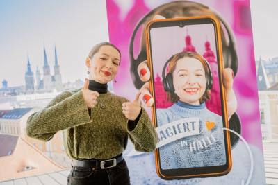 Eine junge Frau steht vor einem Banner, ab dem sie selbst zu sehen ist und ein Handy in der Hand mit der Engagement-App Halle